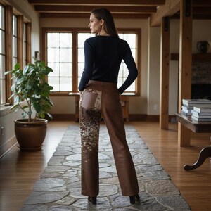 May include: A woman wearing a black long-sleeve shirt and brown leather pants. The pants have a decorative patch with a patterned design. The woman is standing in a room with a stone floor and large windows.