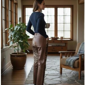 May include: A woman wearing a navy blue long-sleeve shirt and brown leather pants with a patterned design on the back. She is holding a white mug. The setting is a well-lit room with large windows and a stone floor.