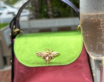 Könnte beinhalten: Eine kleine, farbenfrohe Handtasche mit grünem Deckel und pinkfarbenem Körper, verziert mit einem goldenen Bienenverschluss, steht neben zwei Champagnerflöten, eine gefüllt mit einem sprudelnden Getränk und die andere mit einem gelben Cocktail. Die Szene spielt im Freien.