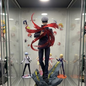 May include: A detailed anime figure of a character in a black outfit with a red translucent energy effect. The figure stands on a rocky base inside a glass display case, with other anime figures and stickers visible.