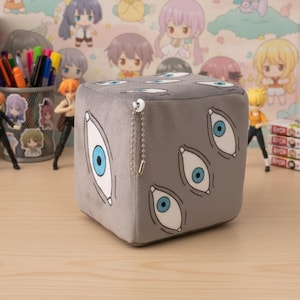 May include: A gray plush cube featuring multiple white-outlined eyes with blue irises. The cube has a small silver chain attached. The background includes anime-style figurines and a colorful wallpaper.