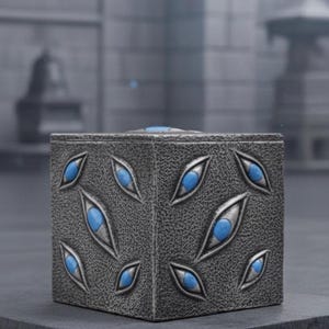 May include: A square, textured, gray box with blue, eye-shaped inlays. The box is set on a dark gray surface, with a blurred background of a building and a pagoda-style structure.