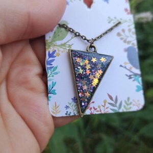 May include: A bronze-toned necklace with a triangular pendant filled with glitter and star-shaped confetti. The pendant is attached to a delicate chain and is displayed on a floral-patterned card.