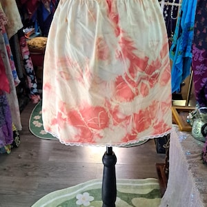 May include: A short, cream-coloured skirt featuring a vibrant coral-red tie-dye pattern throughout. The garment has an elasticated waistband and a delicate white lace trim along the bottom hem, displayed on a black mannequin torso.