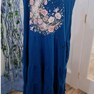 May include: A sleeveless blue tiered dress featuring a graphic print on the upper back. The print displays a crescent moon surrounded by various flowers in shades of pink, white, and purple. The dress has a ruffled hemline with small tassels.