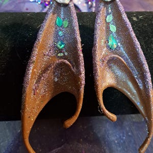 May include: A pair of brown elf ears with glitter and green gem accents. Each ear has a small, light-colored flower at the top. The ears are designed to be worn and have a curved shape.