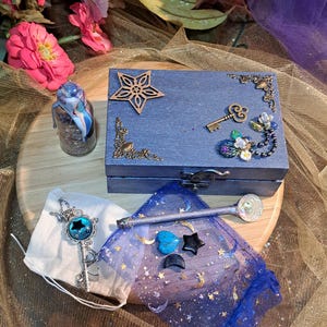 May include: A blue wooden box with decorative metal corners, a star, and a key. A small glass bottle with a blue and white design, a key pendant, a spoon, and small gemstones are also present. The items are arranged on a wooden surface.