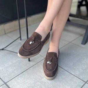 May include: Brown suede loafers with a silver tassel detail. The shoes have a rounded toe and a low, beige sole. The loafers are a slip-on style.