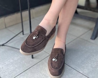 May include: Brown suede loafers with a silver-colored tassel detail. The shoes have a rounded toe and a low, beige sole. The loafers are a slip-on style.