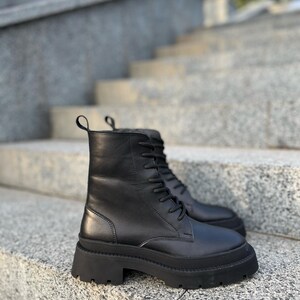May include: A pair of black leather combat boots with a lace-up design. The boots have a chunky, ridged sole and a pull tab at the top. The boots are displayed on a set of gray stone steps.