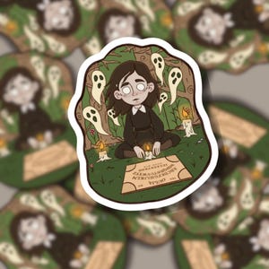 May include: A sticker depicting a person in a black dress, surrounded by ghosts and candles, with a Ouija board. The design has a spooky, mystical theme, with a forest background and the word "Ouija" visible on the board.