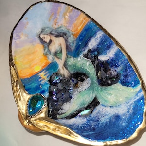 May include: An oyster shell trinket dish featuring a mermaid illustration. The artwork depicts a mermaid with flowing blue hair, sitting on a rock in the ocean. The dish has a gold-colored rim and a blue gemstone accent.