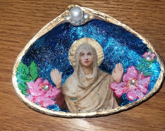 Puede incluir: Arte de concha pintada a mano que representa una figura religiosa con un halo dorado, sobre un fondo azul brillante. Flores rosas y una perla adornan el borde de la concha. La concha está bordeada con pintura dorada.