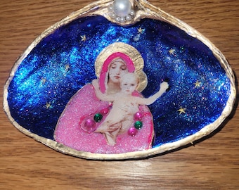 Puede incluir: Concha decorativa con una imagen religiosa de una mujer y un niño. El interior de la concha está pintado con un fondo azul estrellado y un borde dorado. La imagen está acentuada con adornos rosas y verdes y una perla.