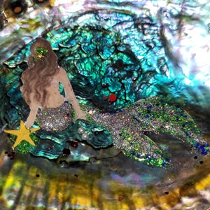 May include: A mermaid figure with glittery green and silver tail and a yellow starfish, set against an iridescent, textured background of blues, greens, and purples. The mermaid has long brown hair and is adorned with glitter.
