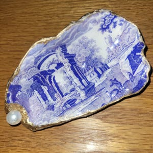 May include: An oyster shell dish with a blue and white landscape design. The shell's edge is trimmed with gold paint, and a pearl is attached to one end. The dish is on a wooden surface.