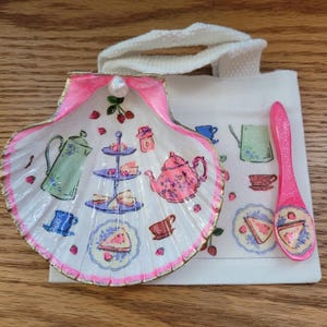 Könnte beinhalten: Dekorative Muschel mit rosa Rand und Perle, die eine Teeparty-Szene mit Teekanne, Kuchenständer und Tassen zeigt. Eine kleine Tragetasche und ein rosa Löffel mit passenden Designs vervollständigen das Set.