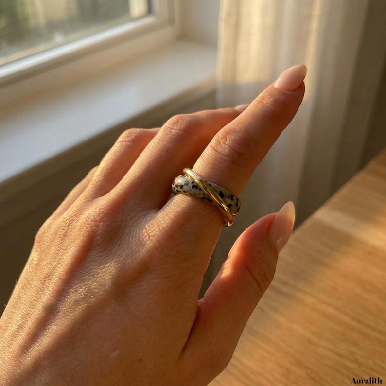 May include: A close-up of a ring with a speckled, gray band and a gold band wrapped around it. The ring is on a person's finger, with the word "Auralith" in the bottom right corner.