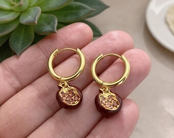 May include: Gold-colored hoop earrings with a pomegranate charm. The charm is a dark red color with small, orange-colored accents. The earrings are shown against a blurred background.