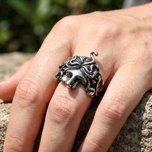 May include: A silver-colored ring featuring a detailed Medusa head design. The ring has a textured surface with intricate curls and a red gemstone eye. The Medusa ring is a unique piece of jewelry.