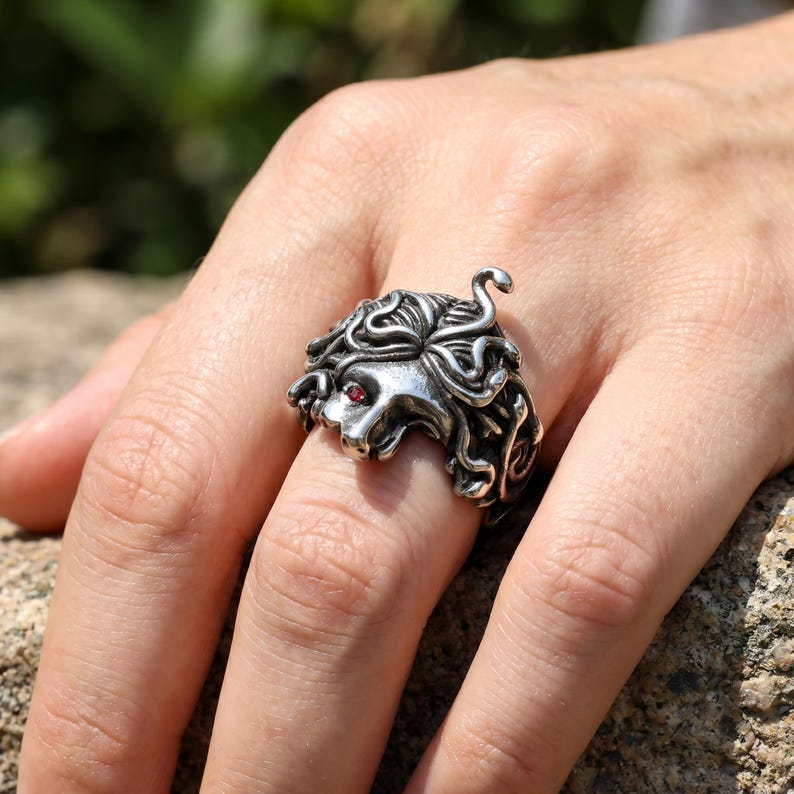 May include: A silver-colored ring featuring a detailed Medusa head design. The ring has a textured surface with intricate curls and a red gemstone eye. The Medusa ring is a unique piece of jewelry.
