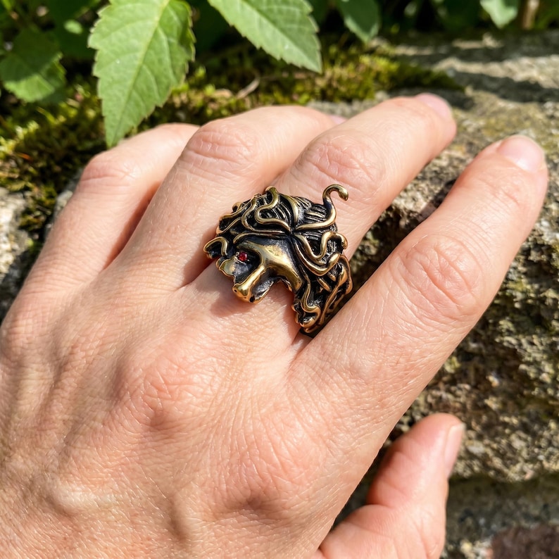 May include: A bronze-toned ring featuring a Medusa head design with intricate details. The ring has a red gemstone eye and a textured surface, showcasing a unique and detailed piece of jewelry.