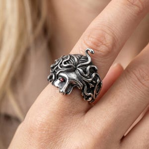 May include: A silver-colored ring featuring a Medusa head design. The ring has intricate details, including curly hair and a red gemstone eye. The snake-like hair is sculpted around the head, creating a unique and detailed piece of jewelry.