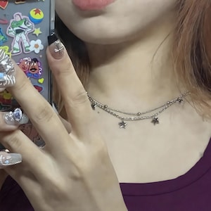 Può includere: Una collana argentata a due fili con ciondoli a forma di stella. La collana ha due fili, uno con piccole perline e l'altro con stelle pendenti. La persona indossa un top viola scuro.