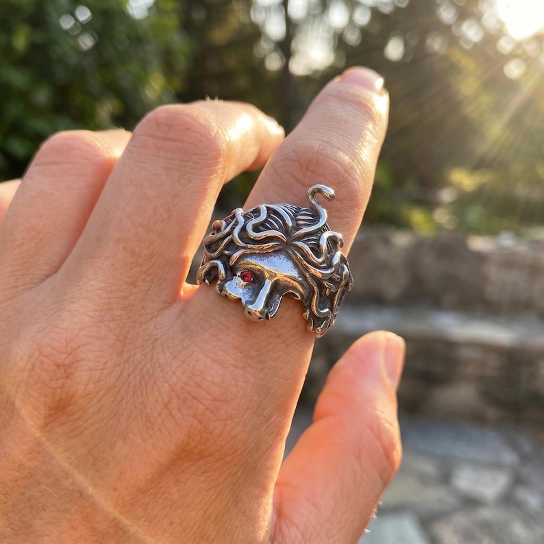 May include: A silver-colored ring featuring a Medusa design. The ring has intricate details, including flowing hair and a red gemstone eye. The ring is worn on a finger, with a blurred background of greenery and sunlight.