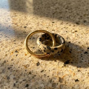 May include: Two rings, one gold-colored and one speckled with black spots on a cream background, interlocked. The rings are set on a speckled, light-brown surface, with shadows cast across the scene. The rings are likely jewelry.