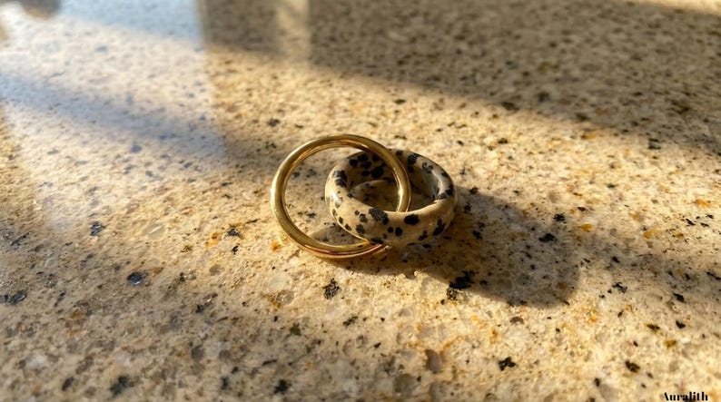May include: Two rings, one gold-colored and one speckled with black spots on a cream background, interlocked. The rings are set on a speckled, light-brown surface, with shadows cast across the scene. The rings are likely jewelry.