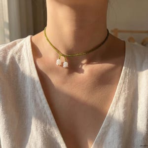 Op de afbeelding: Een delicate ketting met een groene kralenketting en twee witte lelietjes-van-de-vallei-hangers. De ketting wordt om de nek gedragen en toont het bloemendesign. De ketting is een sieraad.