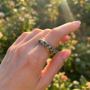 May include: A gold ring with a cylindrical, speckled stone band. The stone is a light beige color with black spots. The ring is shown on a finger against a blurred background of green foliage and sunlight.