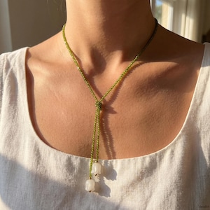 Op de afbeelding: Een delicate groene kralenketting met een Y-vormig ontwerp. De ketting heeft twee witte, bloemvormige hangers met goudkleurige accenten. De ketting wordt gedragen over een witte top.