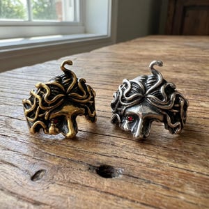 May include: Two Medusa rings, one gold-toned and one silver-toned, each with a red gem eye. The rings feature intricate detailing of Medusa's hair and facial features. The rings are displayed on a wooden surface.