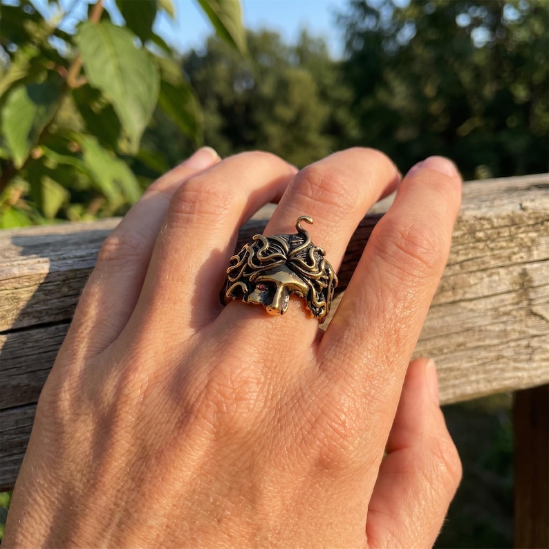 May include: A bronze-colored ring featuring a detailed Medusa design. The ring showcases Medusa's face with intricate curly hair and a snake detail. The ring is a unique piece of jewelry.