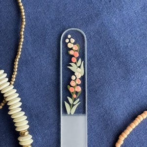 May include: A clear glass nail file with a floral design featuring pink and white flowers and green leaves. The file is rectangular with a rounded top. The background is a textured blue fabric.