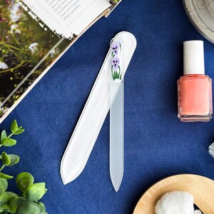 May include: A glass nail file with a white protective sleeve. The file is decorated with purple flowers and green stems. A bottle of coral nail polish is next to the file on a blue surface.