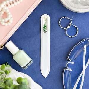 May include: A long, slender glass nail file with a rounded top and pointed bottom, featuring a decorative botanical print of green leaves and small yellow bees at the top end, encased in a white protective sleeve with a clear front window.