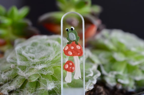Glass nail file hand painted with a small cute green frog sitting on a rust colored mushroom with a off white stem and a smaller mushroom below.