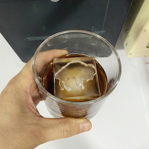 May include: A clear glass of amber-colored liquid with a large ice cube featuring a bison design. The ice cube is partially melted, and the glass is held by a hand. The drink appears to be whiskey or a similar spirit.