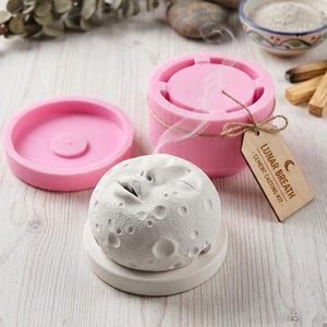 May include: A white, textured moon-shaped cement casting with a face, sitting on a white base. A pink mold and a pink container are nearby, along with a wooden tag that reads "Lunar Breath - Cement Casting Kit".