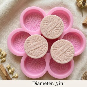 Puede incluir: Un molde de jabón de silicona rosa con seis cavidades circulares, cada una con un diseño de árbol y el texto "NECTAR & ROOTS". Tres jabones beige, de 7.6 cm de diámetro, están en el molde. Los jabones también tienen el texto "NECTAR & ROOTS".