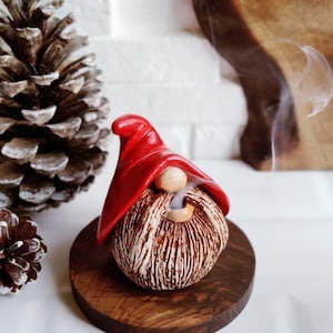 May include: A ceramic gnome figurine with a red hat and a textured, brown and white beard. The gnome is on a small, round, wooden base. Smoke is rising from the gnome's beard, suggesting it is an incense holder. A pine cone is in the background.
