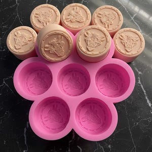 Peut inclure: Ensemble de moules à savon circulaires rose clair avec un motif de tête de vache, ainsi qu'un moule en silicone rose assorti. Les moules à savon sont disposés sur une surface sombre, mettant en valeur les empreintes détaillées de la tête de vache.