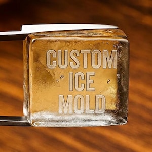 Könnte beinhalten: Ein klarer, quadratischer Eiswürfel, der von einer Metallzange gehalten wird und in der Mitte den eingravierten Text CUSTOM ICE MOLD trägt. Der Eiswürfel hat einen goldbraunen Farbton und sichtbare Luftblasen in seiner Struktur.
