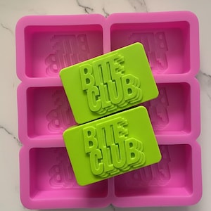 Puede incluir: Un molde de silicona rosa brillante con seis cavidades rectangulares. Dos pastillas de jabón verde lima se colocan dentro del molde, cada una con el texto "BITE CLUB" en relieve. El molde y las pastillas de jabón están sobre una superficie de mármol blanco.