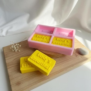 May include: A pink soap mold with four compartments, two filled with yellow soap bars. The bars are embossed with the words "Lady's Essential". Two additional yellow soap bars are in front of the mold. The items are on a wooden board.