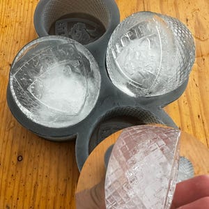 May include: A dark gray silicone ice mold with four spherical cavities, each holding a clear ice sphere. The ice spheres have a textured surface and the word "FLORIDA" is visible on some. A partial ice sphere is held in a hand.