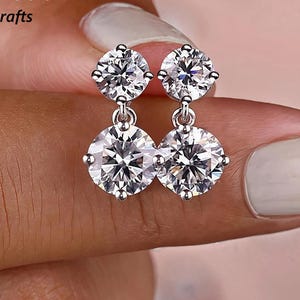 May include: A pair of silver-toned drop earrings featuring two clear, round-cut gemstones. The earrings are held by a person with a light skin tone. The gemstones have multiple facets, reflecting light. The earrings are a classic jewelry design.